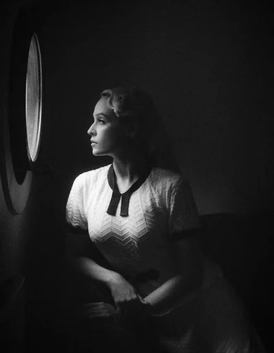 Femme au look années 50, regardant par l’hublot, regard tourné vers la lumière entrant par l’hublot.