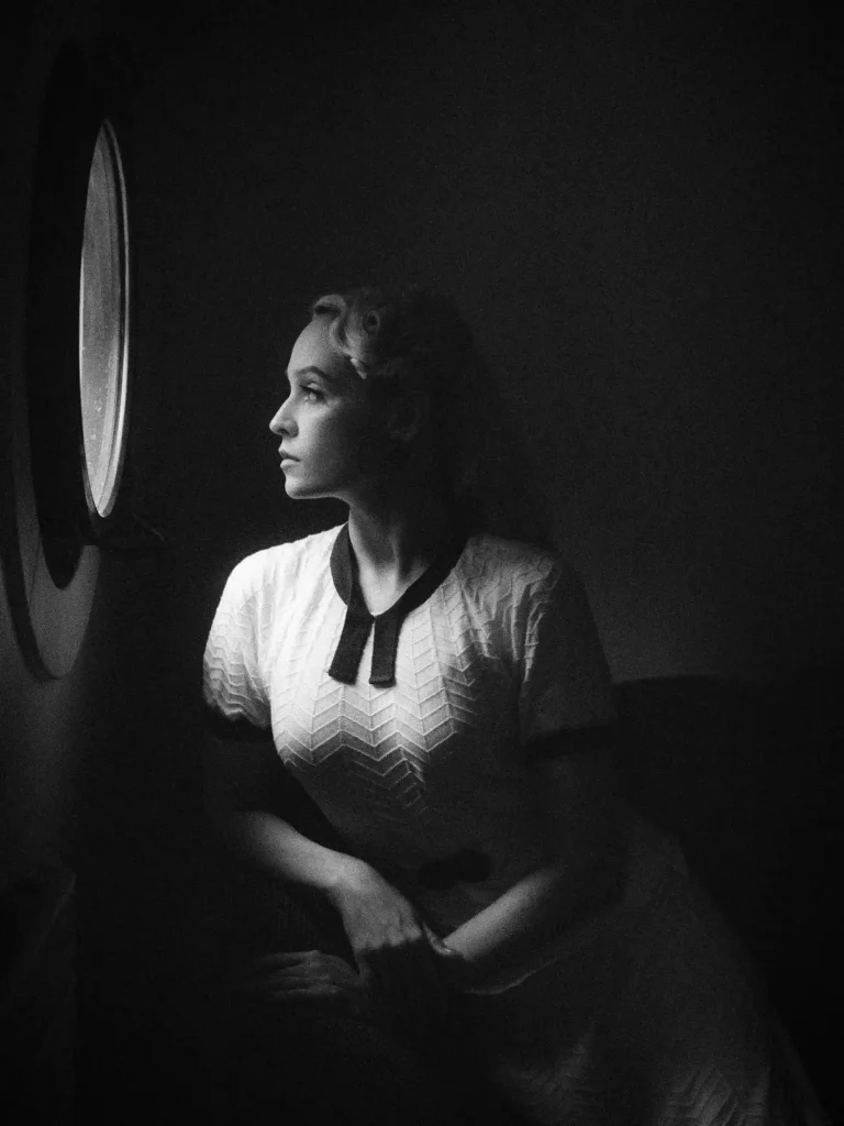 Femme au look années 50, regardant par l’hublot, regard tourné vers la lumière entrant par l’hublot.