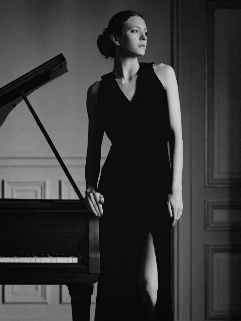 Femme élégante debout, main posée sur un piano, visage tourné sur le côté.