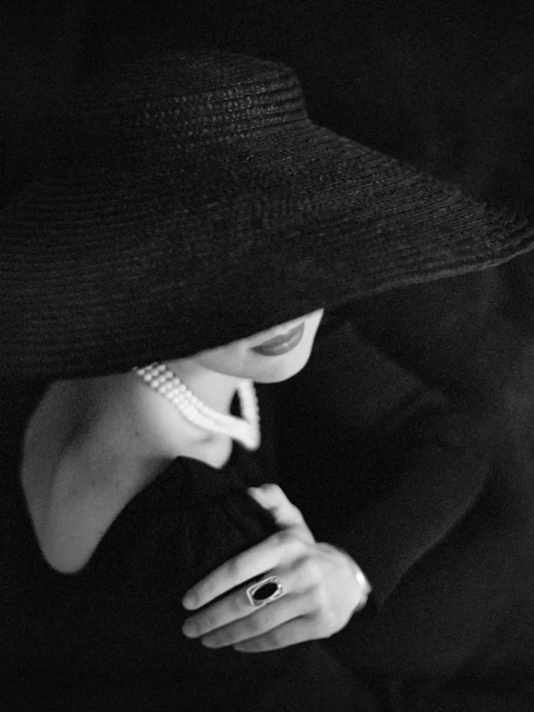 Portrait noir et blanc, femme chic en tenue sombre, chapeau imposant, cadrage partiel sur le visage.